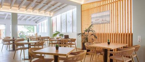 Un amplio comedor interior con mesas y sillas de madera, ventanas grandes y un muro de listones de madera con un cuadro.