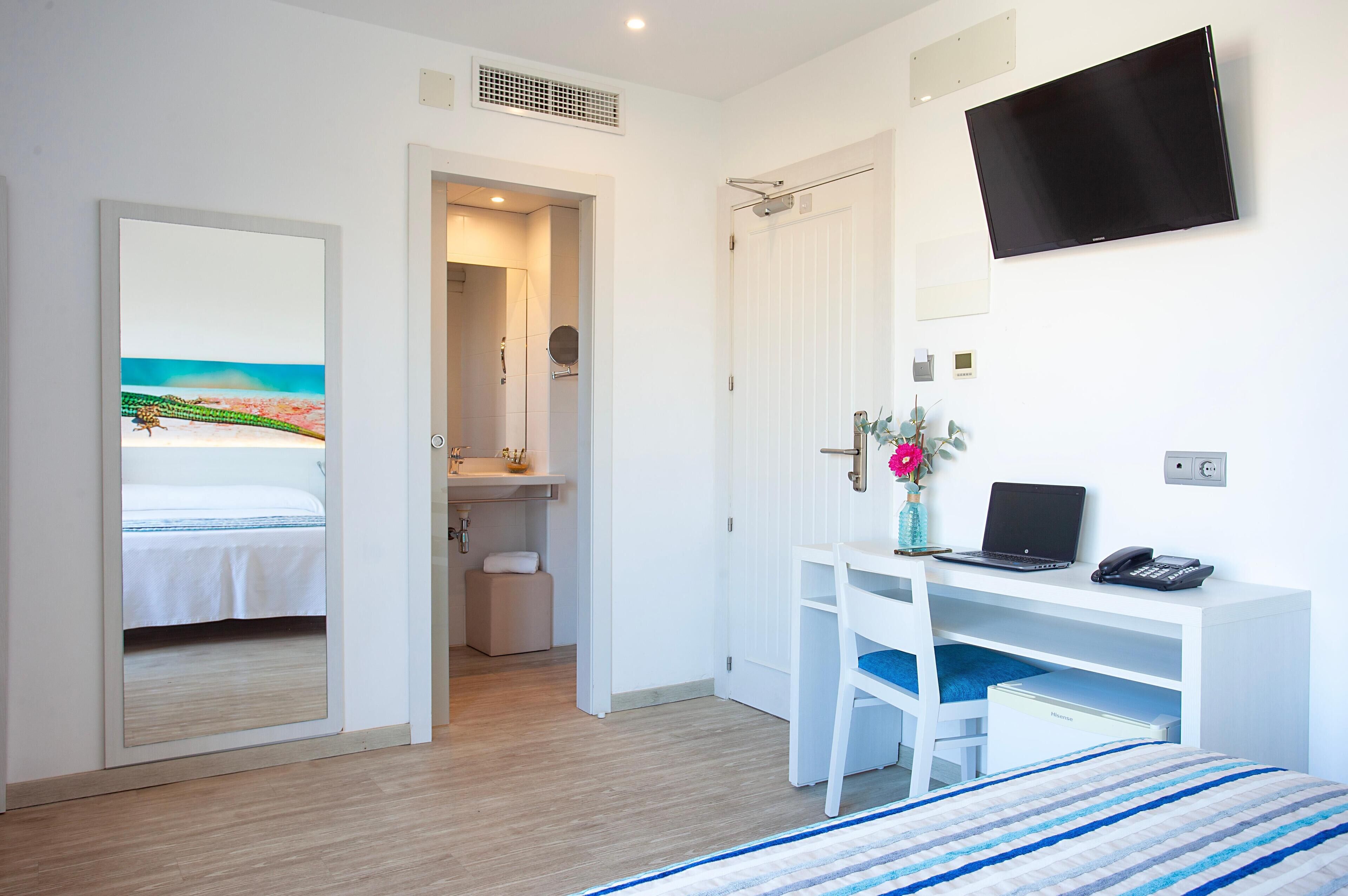 A hotel room with a bed, a desk with a laptop, a TV, and a bathroom visible through an open door.