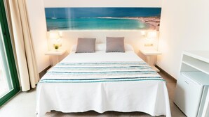 Une chambre d’hôtel avec un lit, deux tables de chevet avec des lampes, une vue sur la plage et une grande fenêtre avec des rideaux.