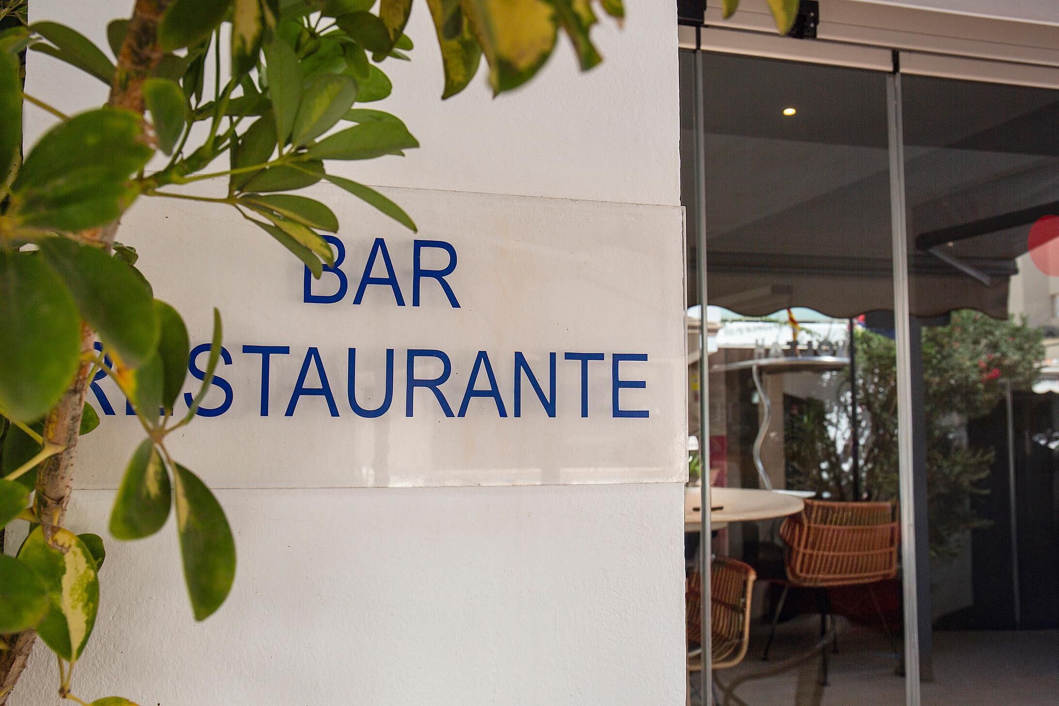 Ein Schild für eine Bar und ein Restaurant, durch Glastüren ist ein Außenbereich mit Sitzplätzen sichtbar.