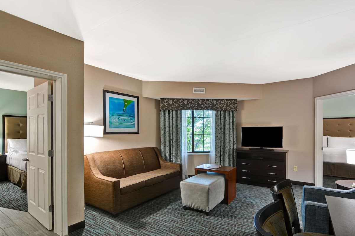 Ein Hotelzimmer mit braunem Sofa, einem kleinen Hocker, einem Fernseher auf einer Kommode und einem Fenster mit Vorhängen.