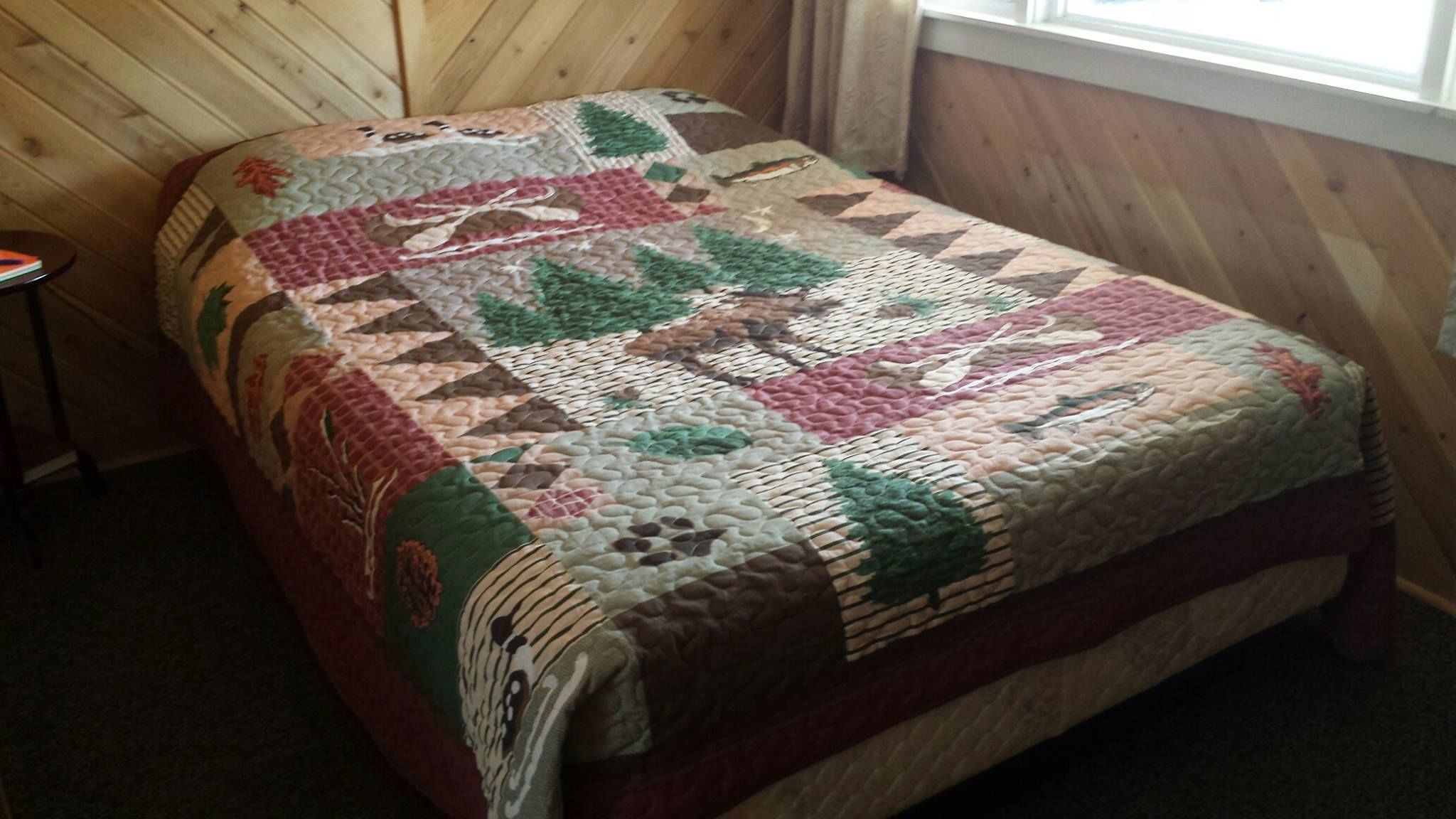 A bed with a quilt featuring a forest and animal pattern, in a room with wooden walls and a window.