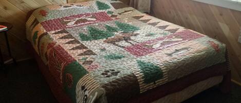 A bed with a quilt featuring a forest and animal pattern, in a room with wooden walls and a window.