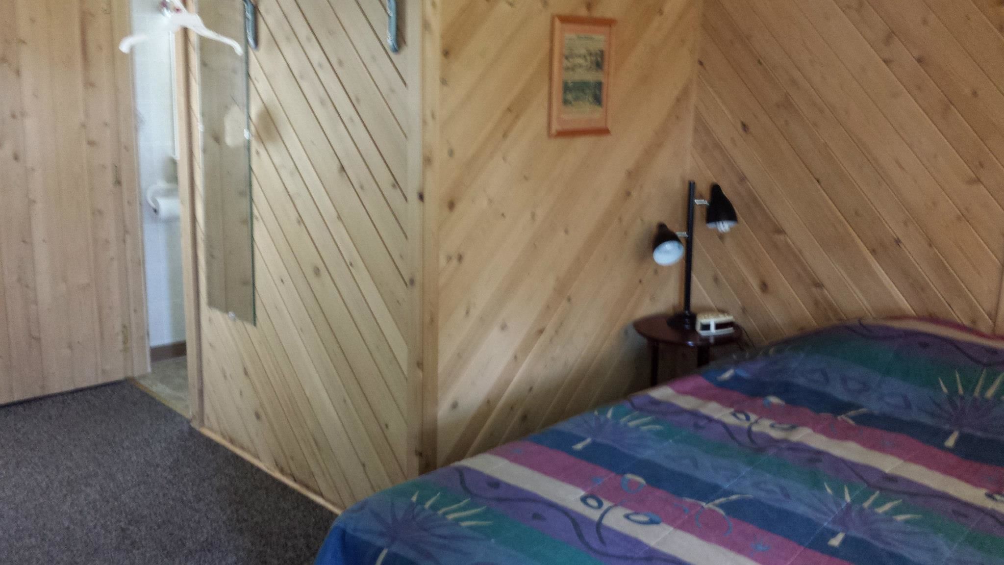 A wooden-paneled room with a bed, a nightstand, a lamp, and a door leading to another room.