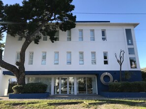 A modern building with a white facade, multiple windows, and a large tree in front.
