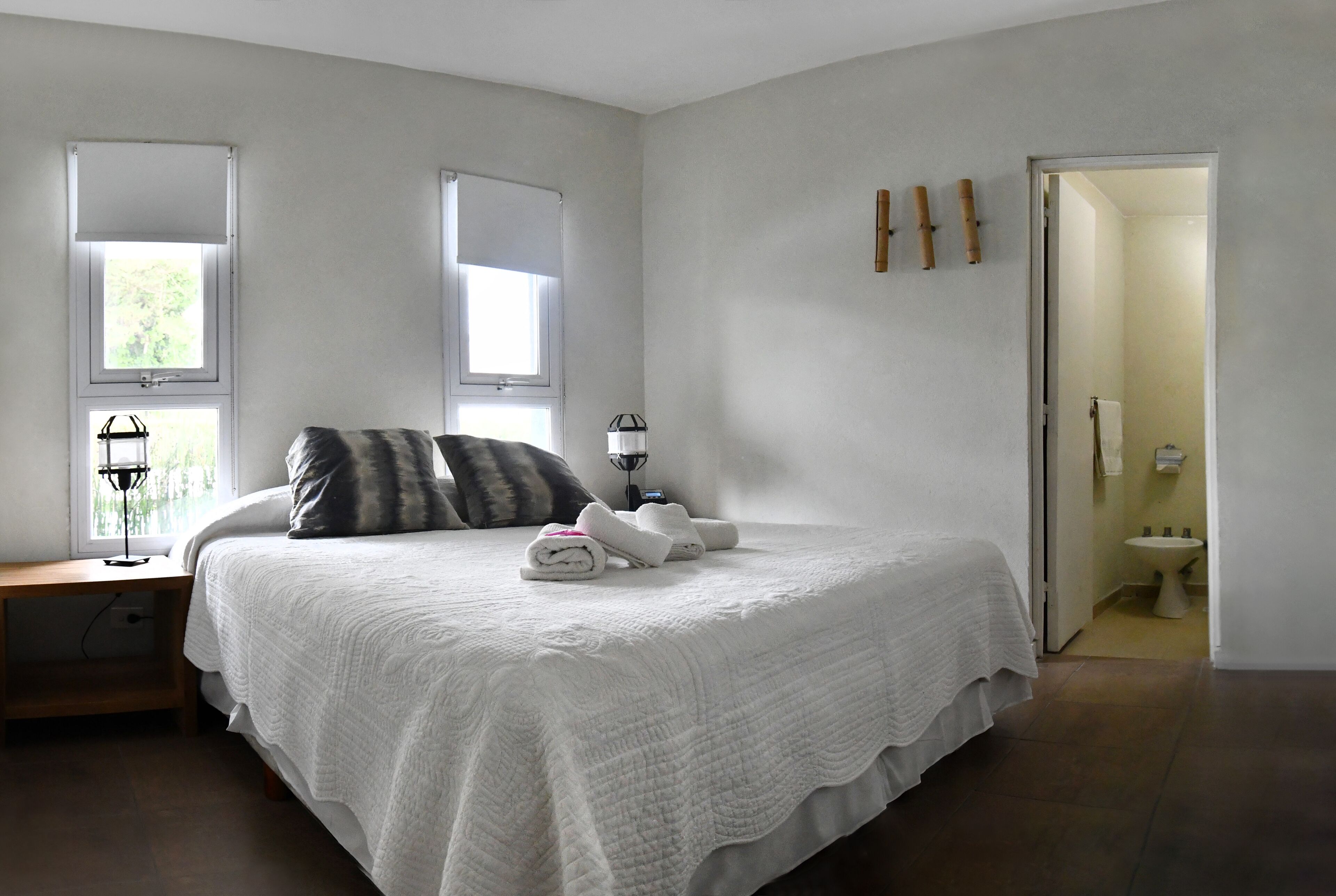 A bedroom with a large bed, two windows, a bedside table, and a door leading to a bathroom.