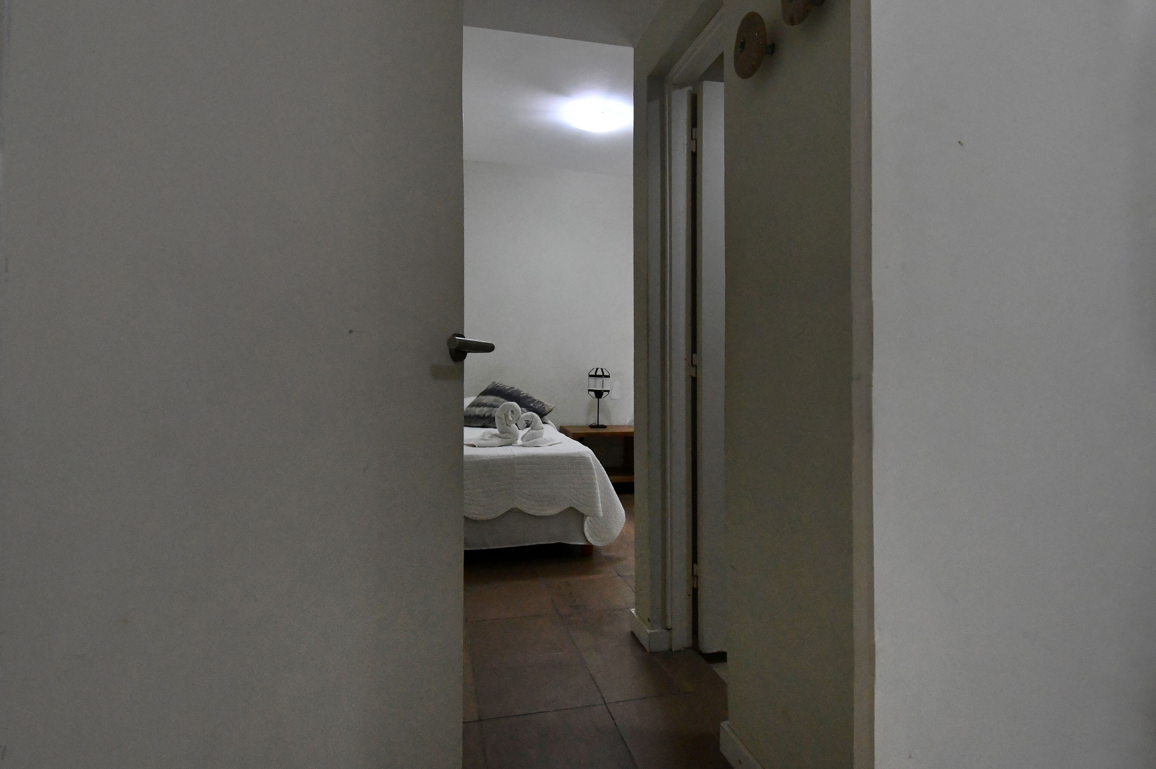 A narrow hallway leading to a bedroom with a bed, a bedside table, and a lamp.