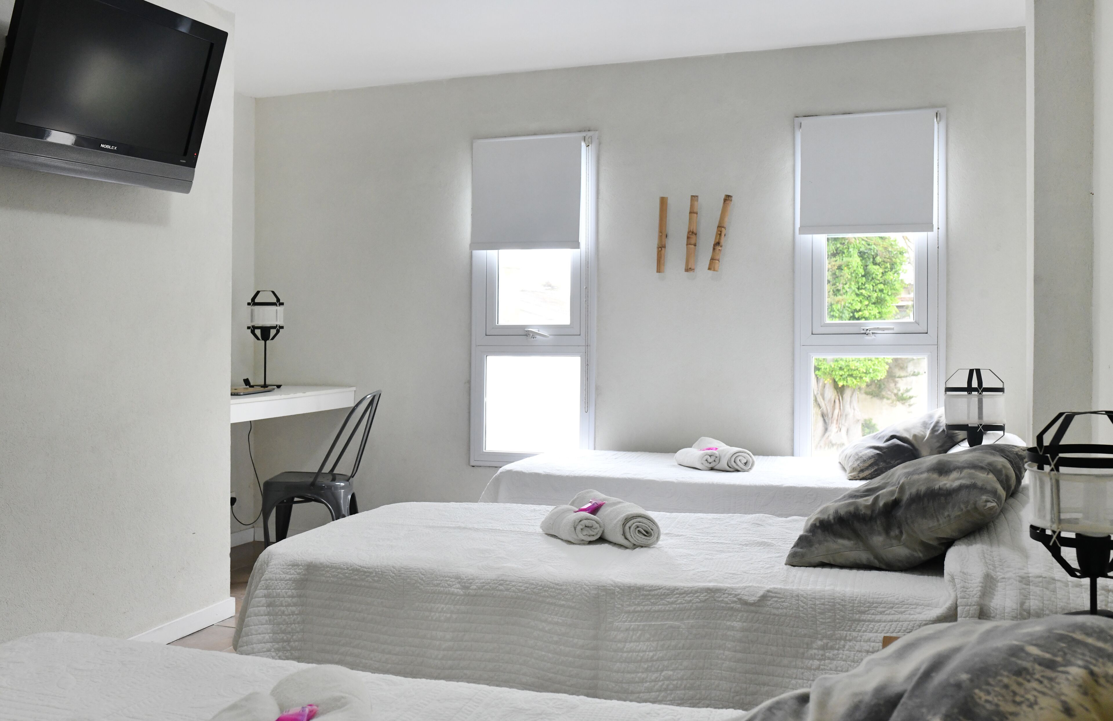 A hotel room with two beds, a desk, a chair, a TV, and two windows with blinds.