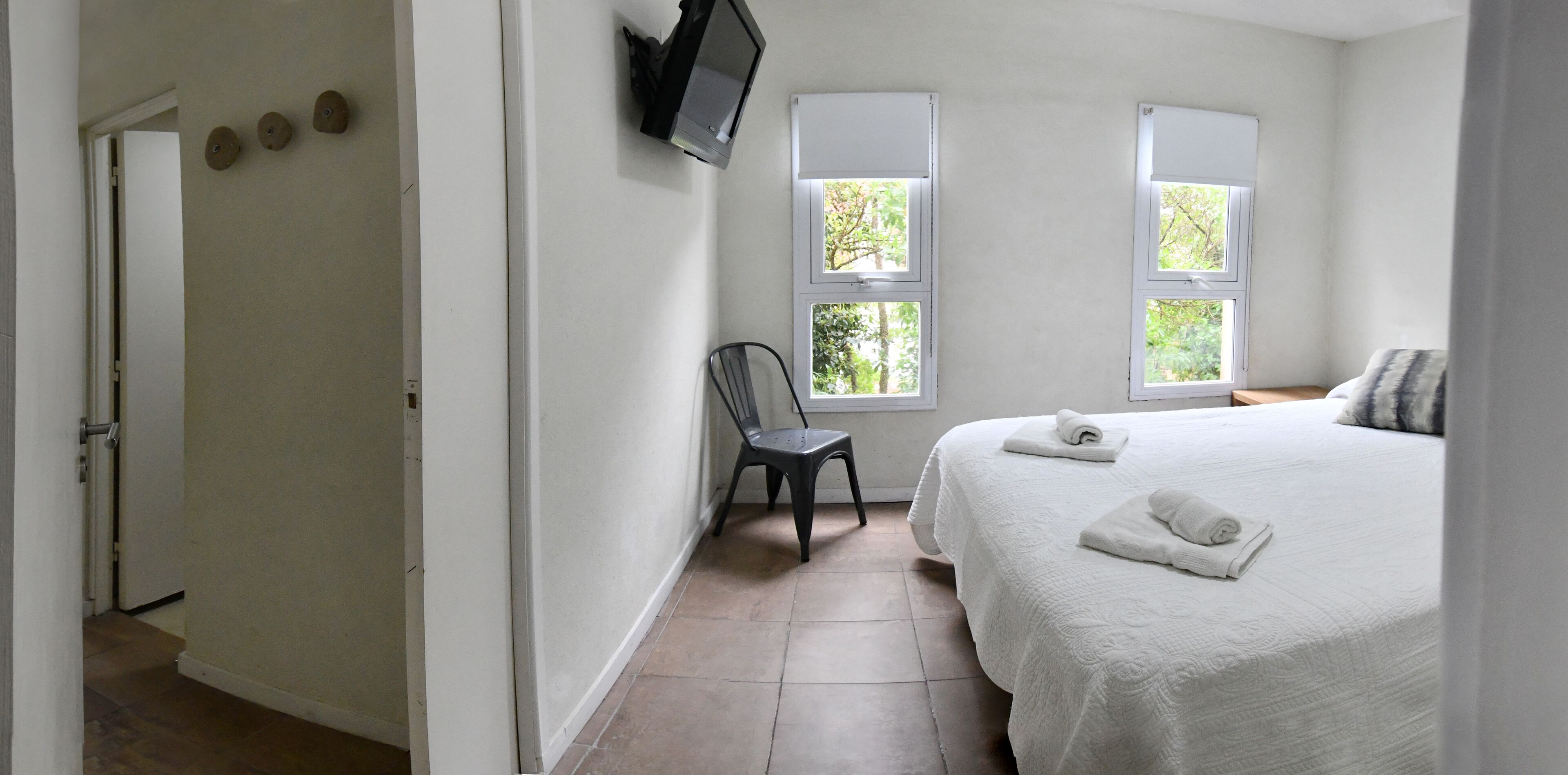 A bedroom with a bed, two windows, a chair, and a TV mounted on the wall.