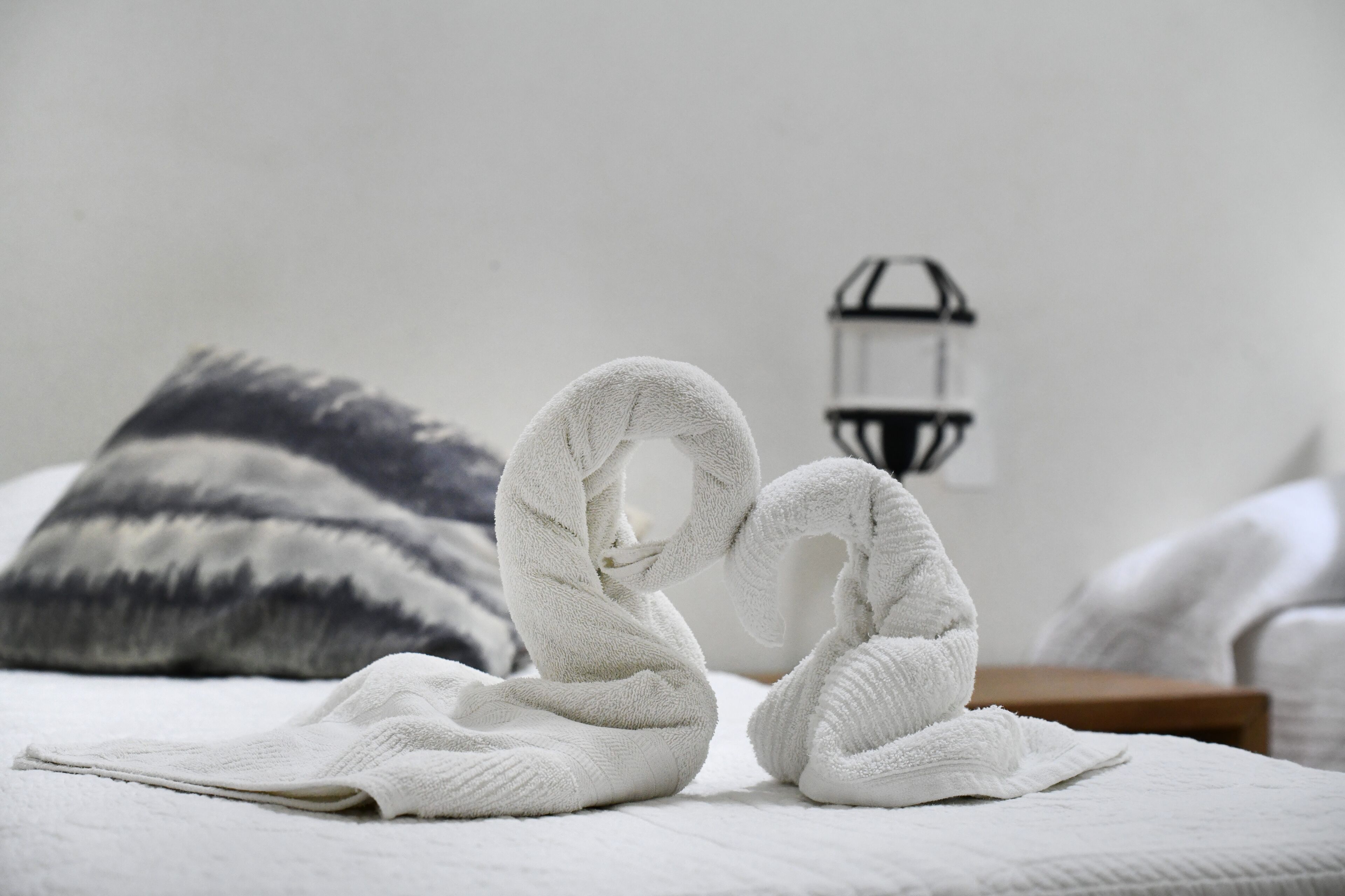 A bed with white towels arranged in a swan shape, a gray and white patterned pillow, and a wall-mounted lantern.