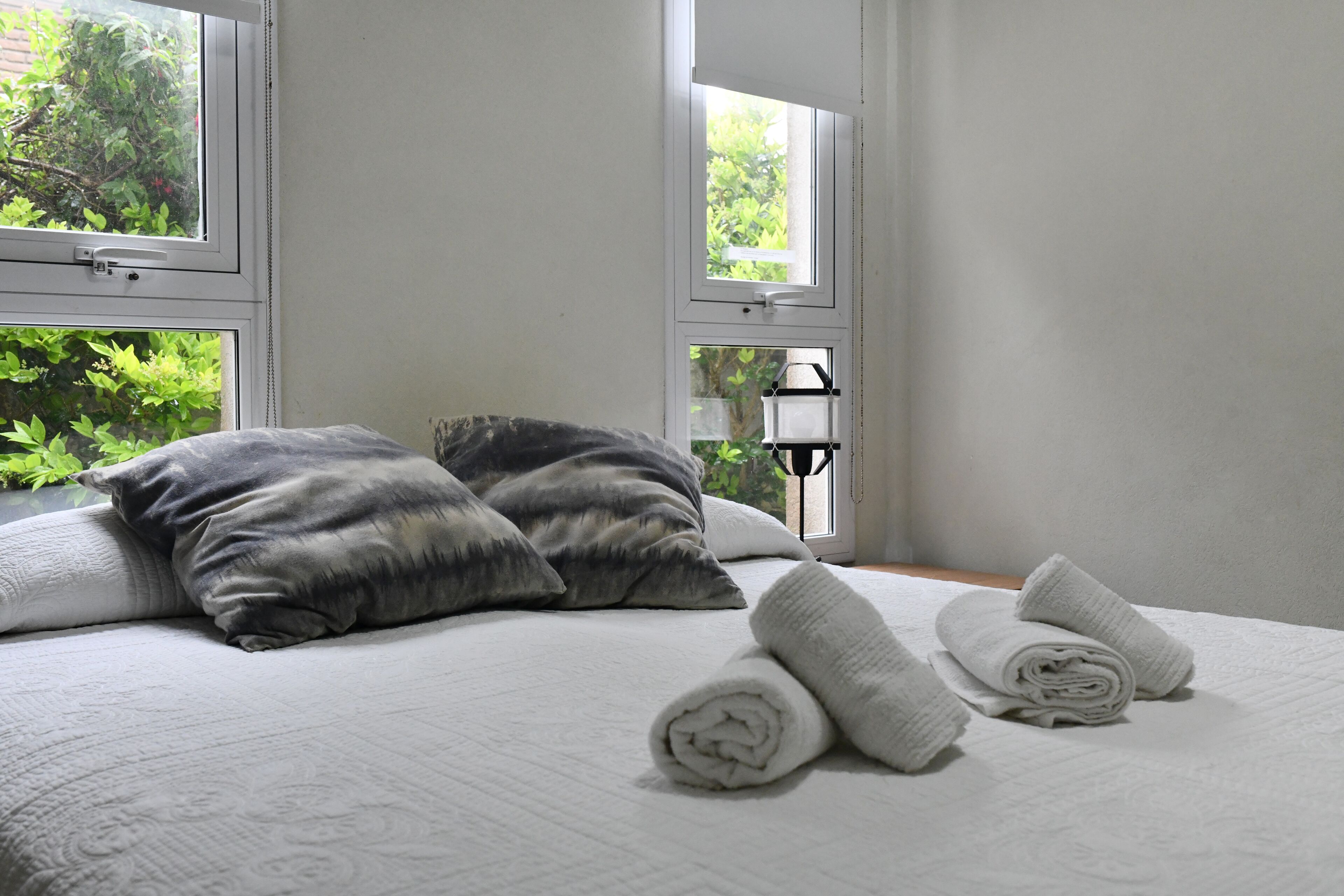 A neatly made bed with pillows and rolled towels, large windows with greenery outside, and a small table with a lamp.