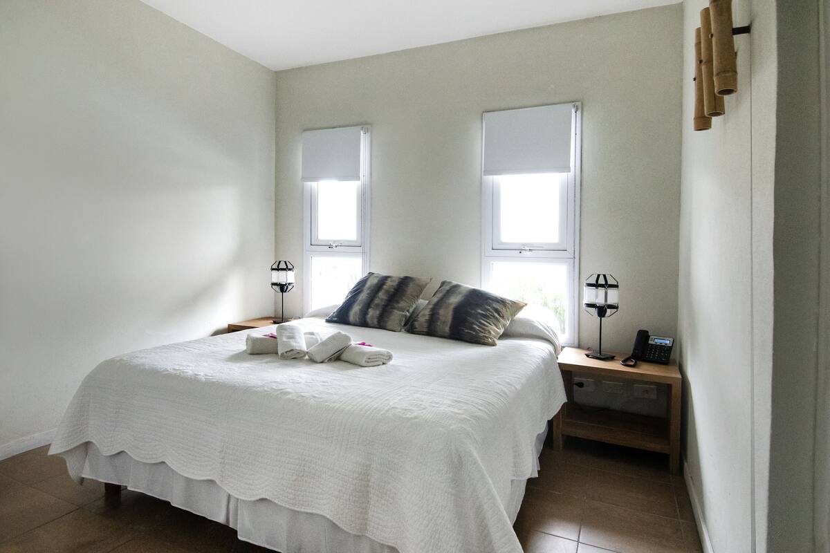 A bedroom with a bed, two bedside tables, a nightstand, and two windows.