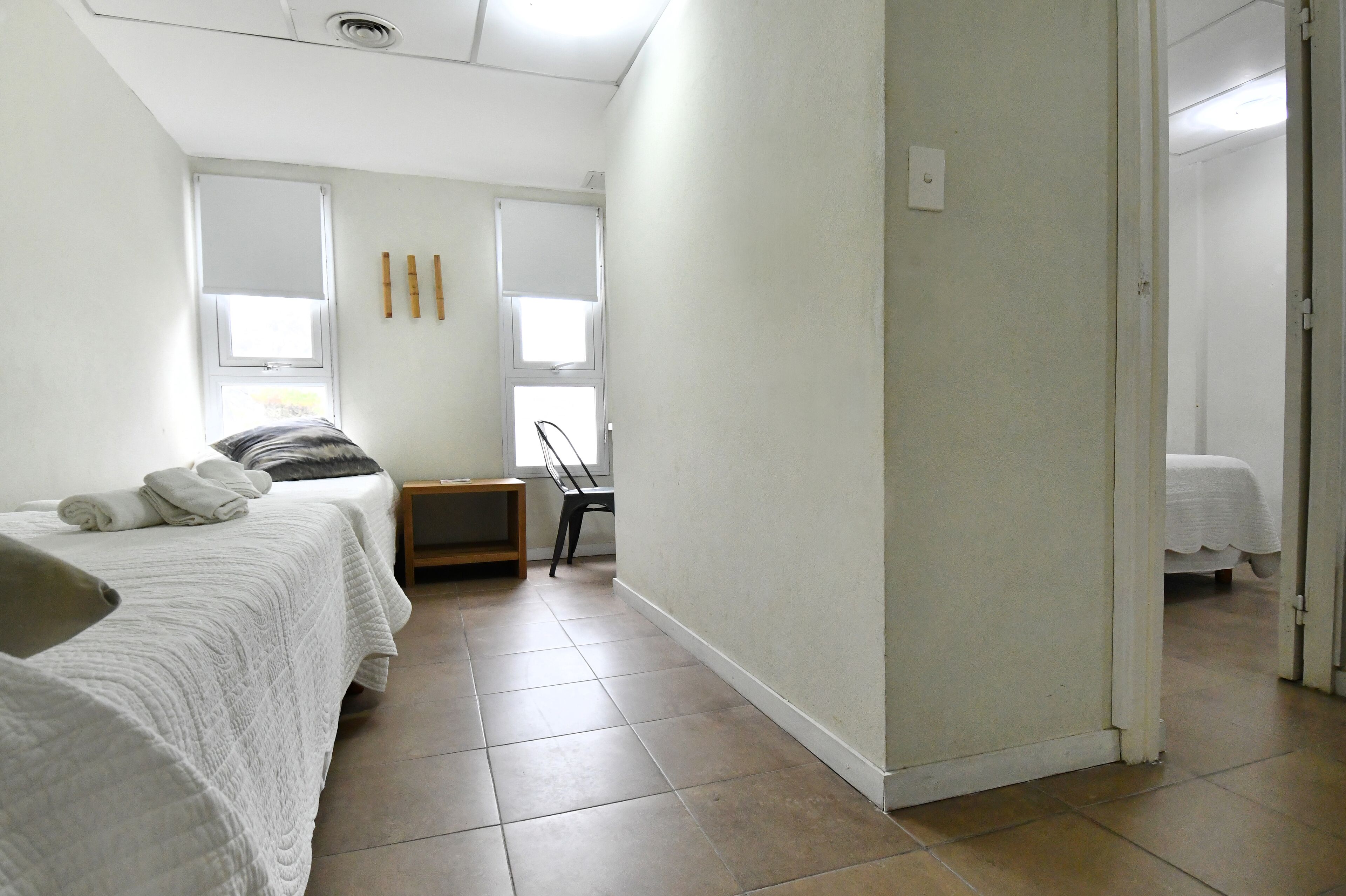 Quarto de hotel com duas camas, uma escrivaninha e uma cadeira. Há duas janelas com persianas, uma porta e piso ladrilhado.