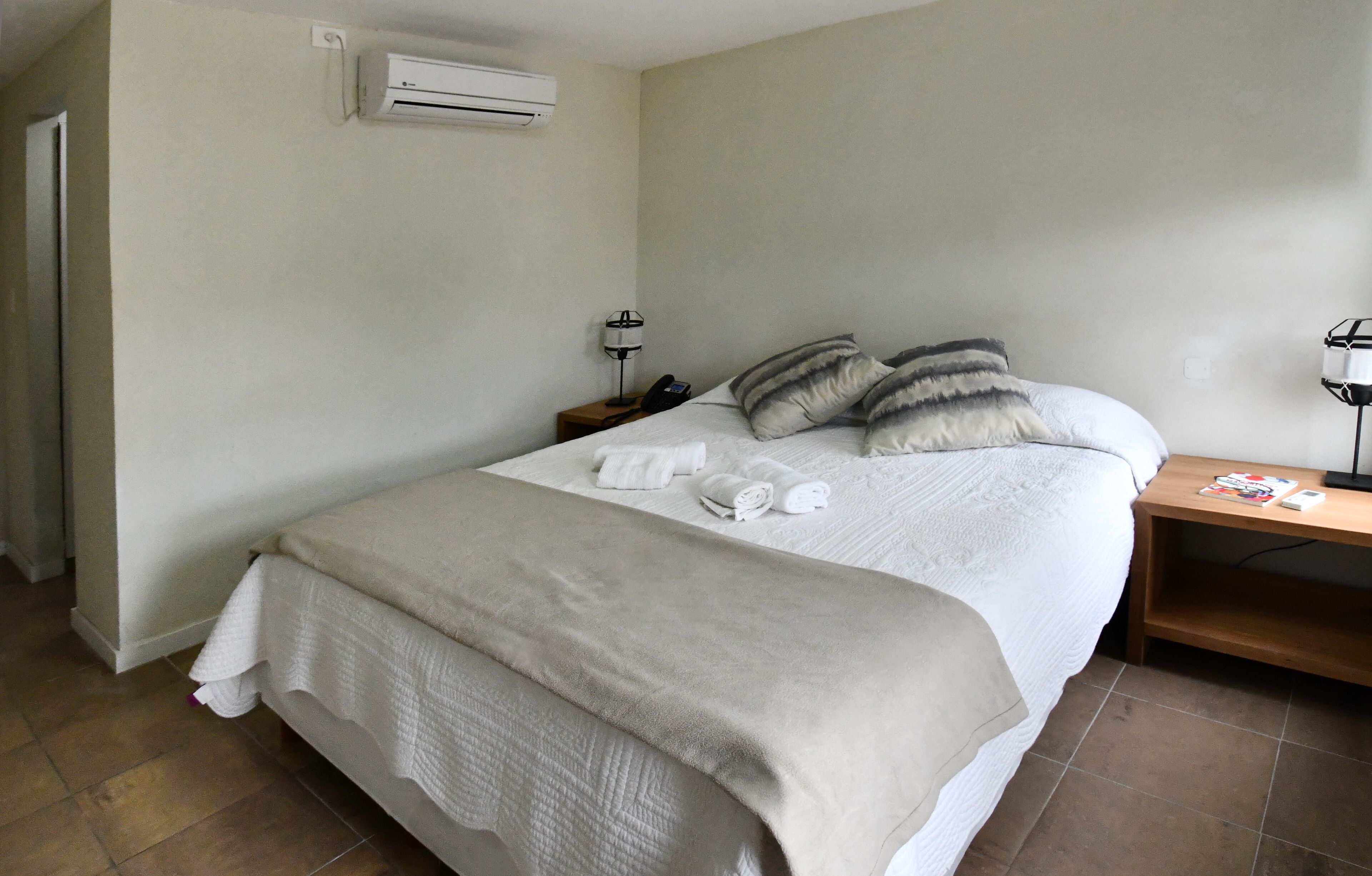 Una cama bien hecha con sábanas blancas y una manta color beige, una mesita de noche con una lámpara y un teléfono, y un equipo de aire acondicionado en la pared.