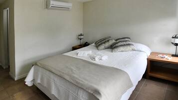 Una cama bien hecha con sábanas blancas y una manta color beige, una mesita de noche con una lámpara y un teléfono, y un equipo de aire acondicionado en la pared.
