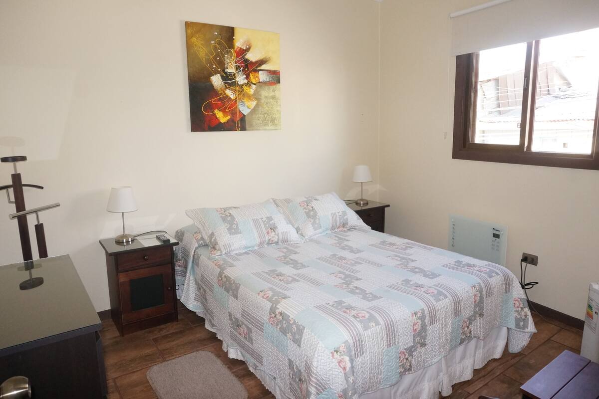 A bedroom with a bed, bedside tables, a desk, a lamp, a radiator, and a window.
