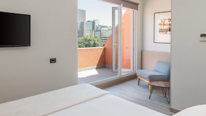 A modern hotel room with a large bed, a seating area with a chair and table, a television, and a sliding glass door leading to an outdoor balcony with a city view. - Novapark (Santiago)