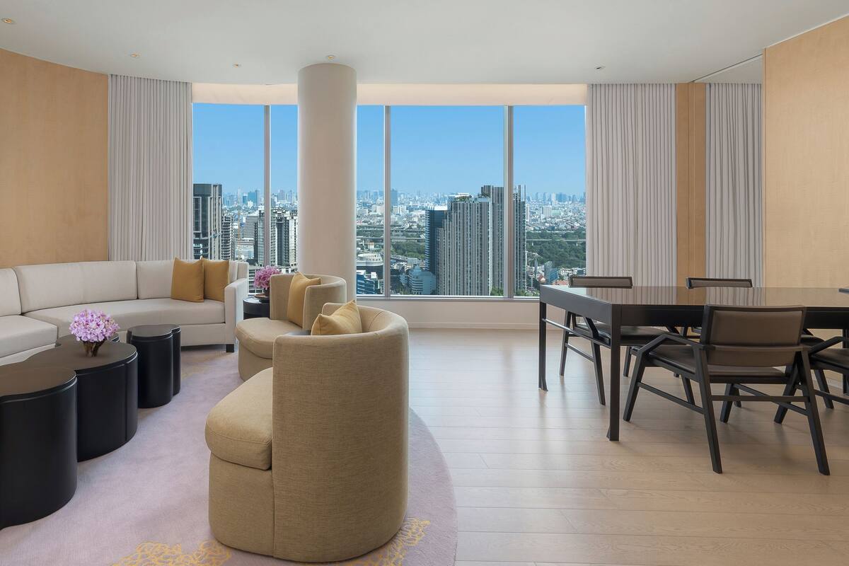 Une chambre d’hôtel moderne avec un canapé incurvé, une table basse ronde, un petit fauteuil, une table à manger avec des chaises et une grande fenêtre donnant sur la ville.