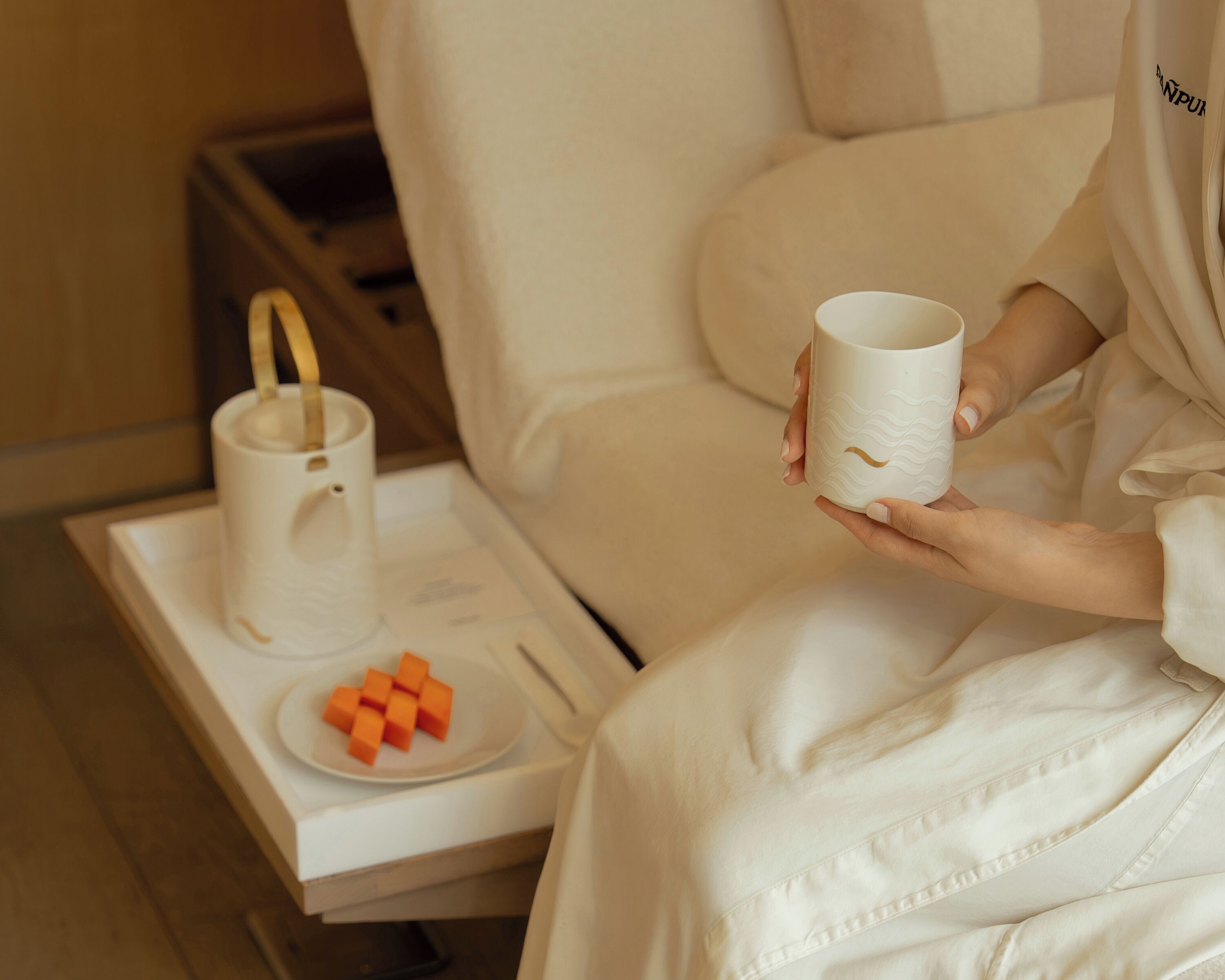 A person in a white robe holding a white mug, sitting on a bed with a tray containing a plate of orange cubes and a white mug.