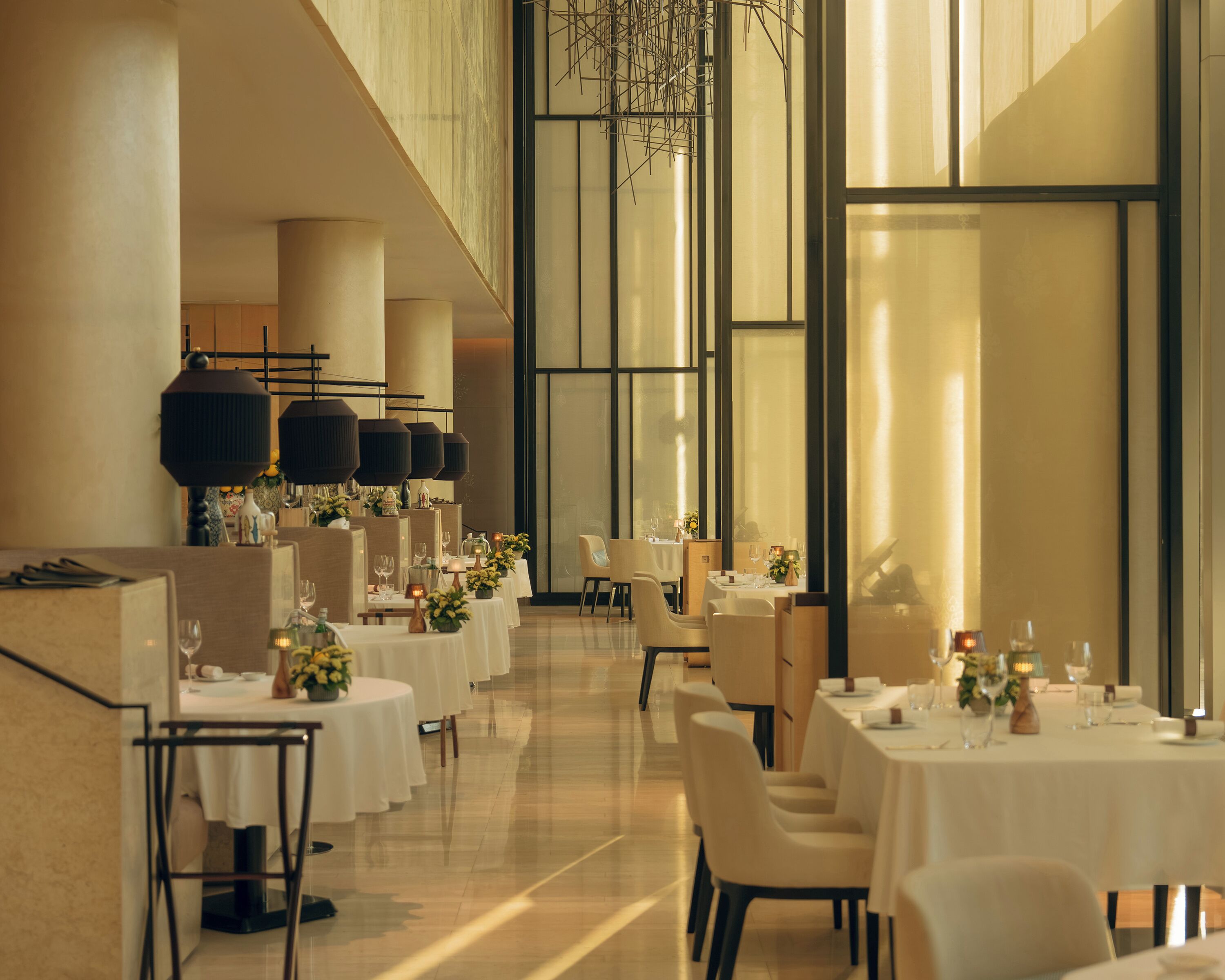 Ampia sala da pranzo con numerosi tavoli e sedie, grandi finestre e lampade dal design moderno.