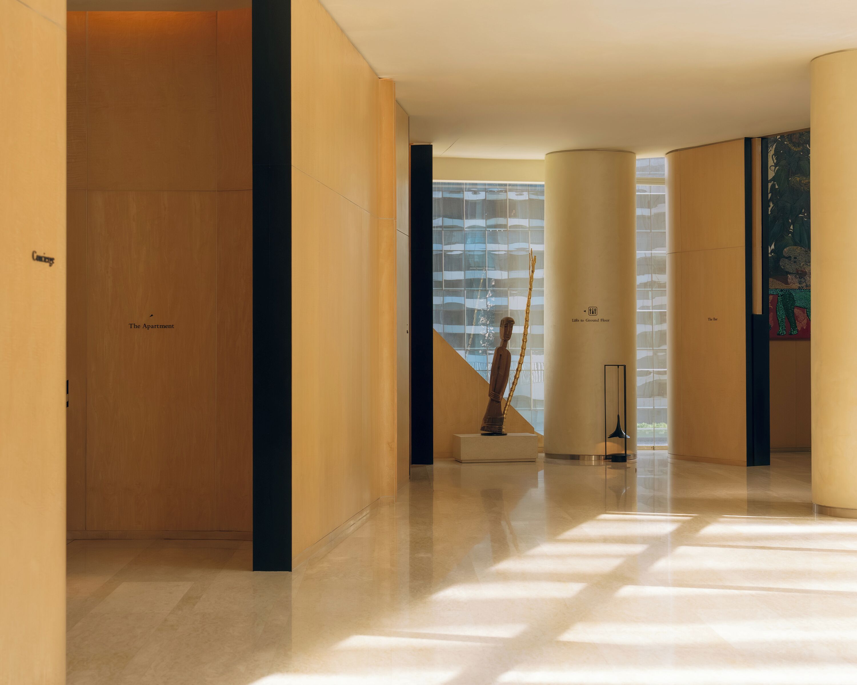 A modern hallway with a wooden door labeled 'The Apartment,' a sculpture, and a staircase leading to a glass building.
