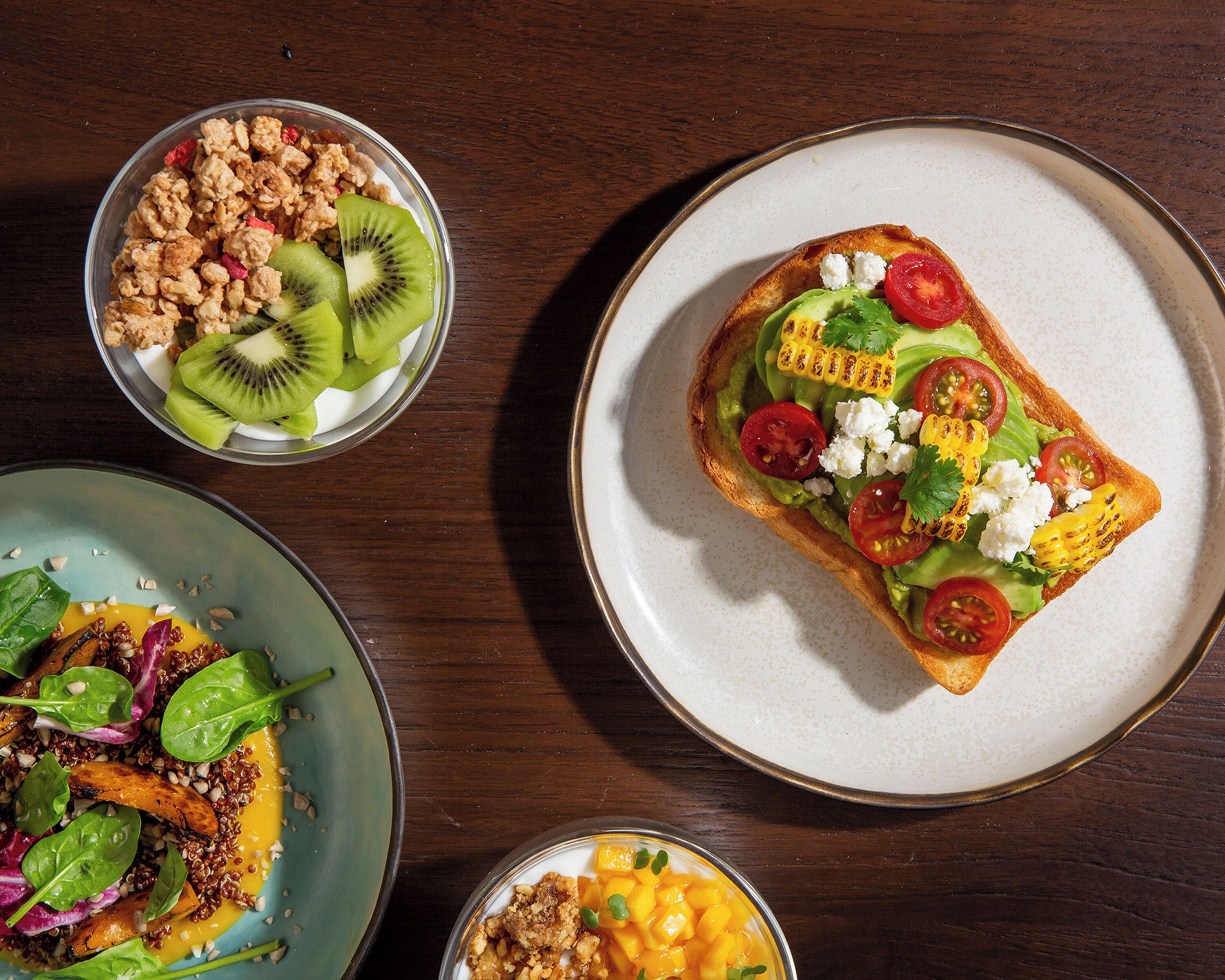 Una mesa con tres platos: una ensalada de espinacas y nueces, un bol de fruta con kiwi y granola, y un sándwich de aguacate, tomate y maíz.