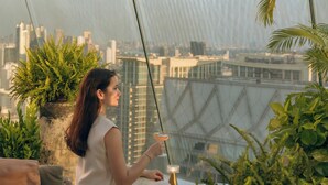 Una persona sentada en un balcón con vista a la ciudad, sosteniendo una bebida, rodeada de plantas y una baranda moderna.