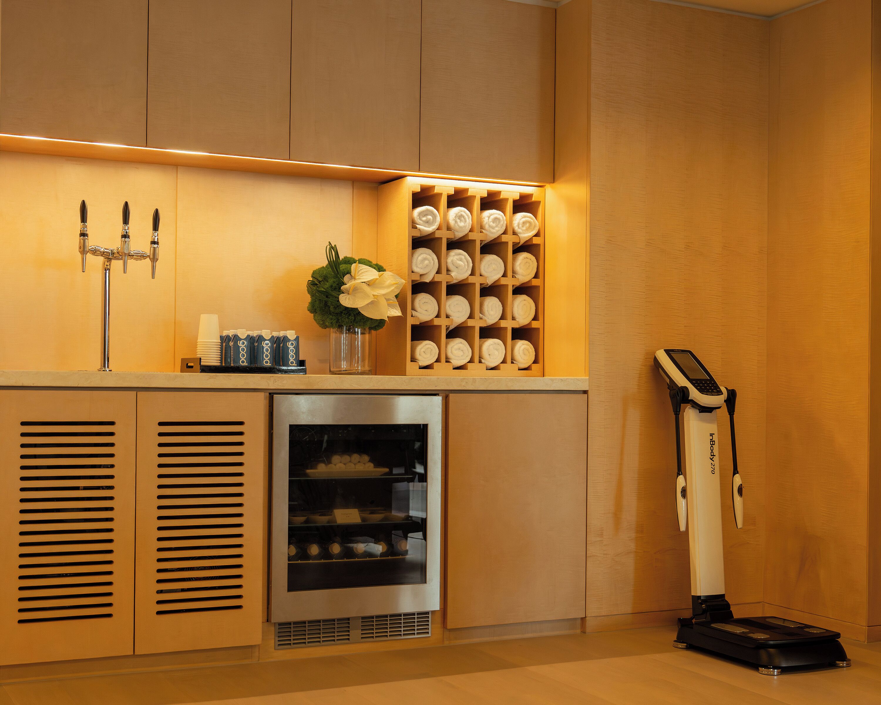A hotel reception area with a counter, a beverage dispenser, a vase with flowers, a towel rack, a refrigerator, and a vacuum cleaner.