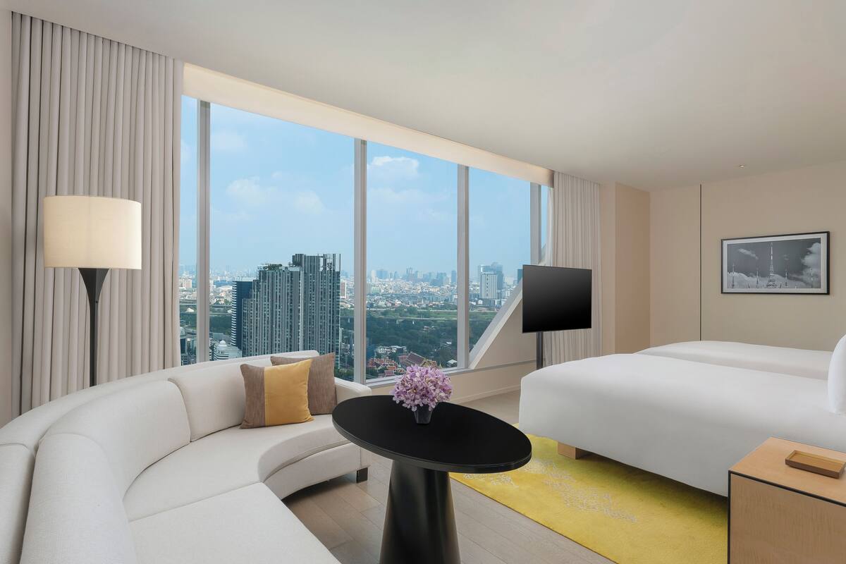 Une chambre d’hôtel moderne avec un canapé incurvé, une petite table surmontée d’un vase de fleurs, un lit, une télévision et une grande fenêtre donnant sur la ville.