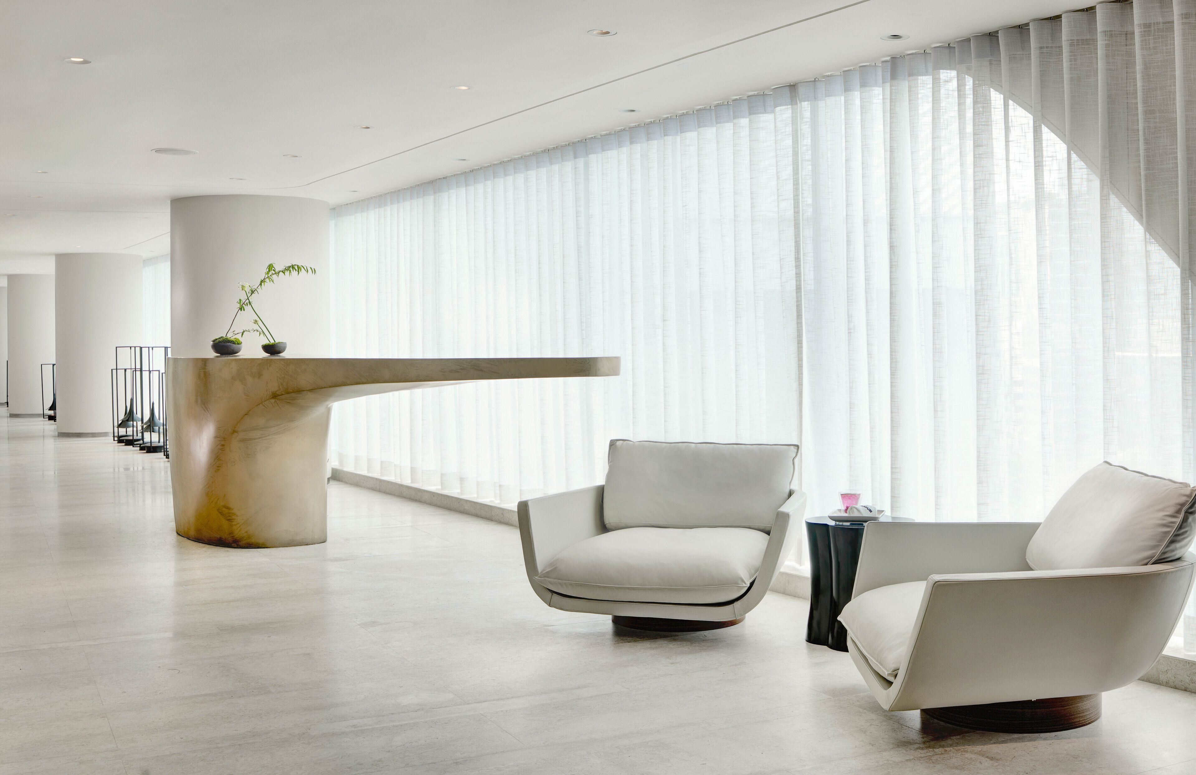 Een moderne hotellobby met een receptiebalie, twee fauteuils en een groot raam met dunne gordijnen.
