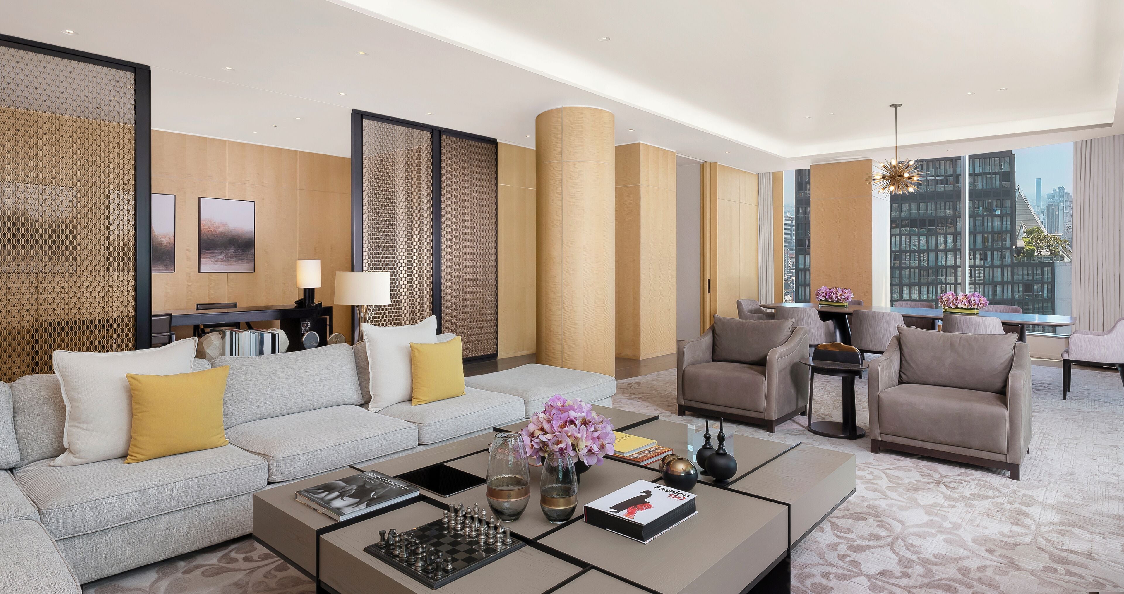 A modern hotel room with a sofa, armchairs, a coffee table, and a view of the city.