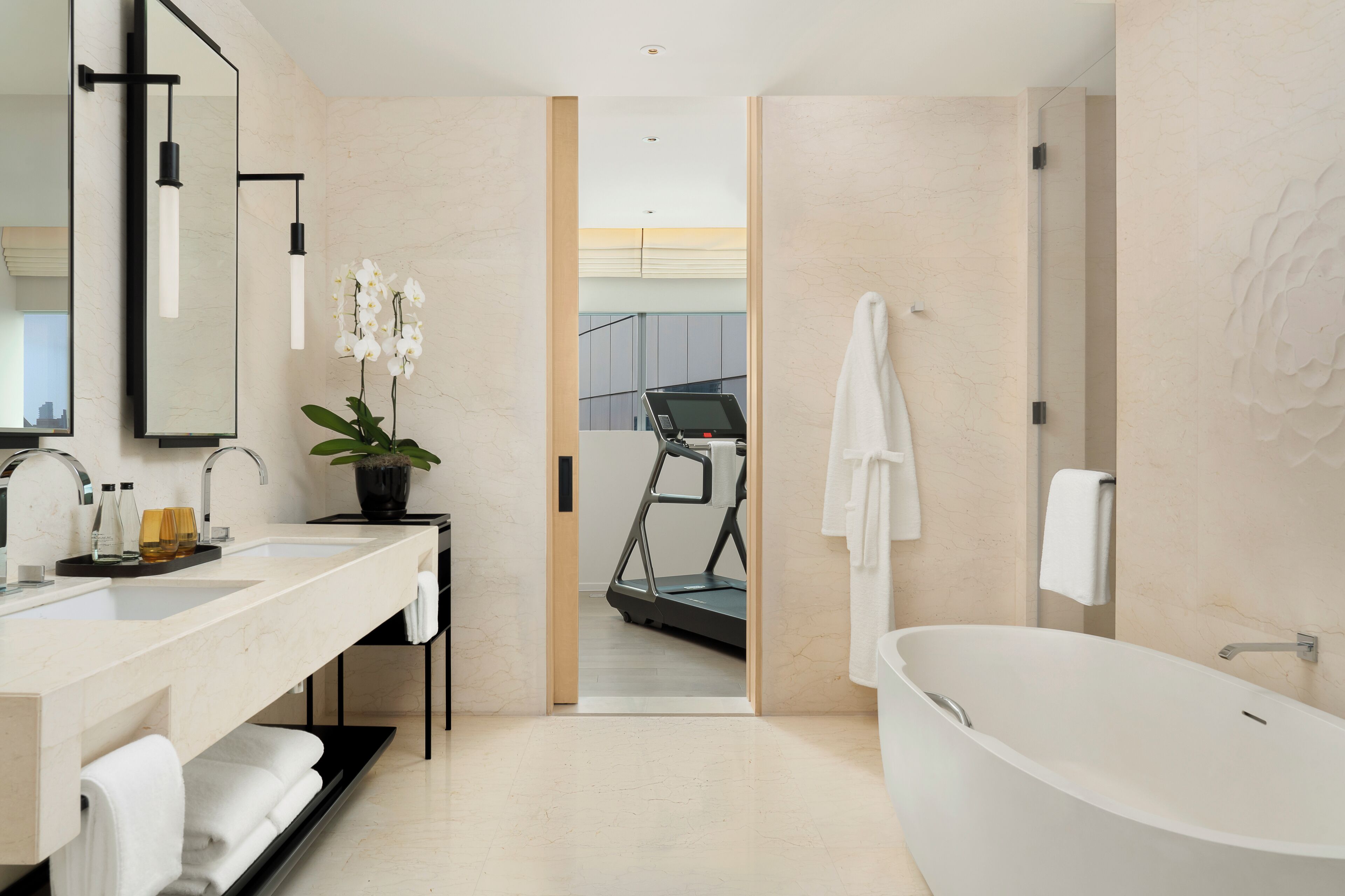 A modern bathroom with a large bathtub, a double sink vanity, a treadmill in the background, and a potted orchid on the counter.