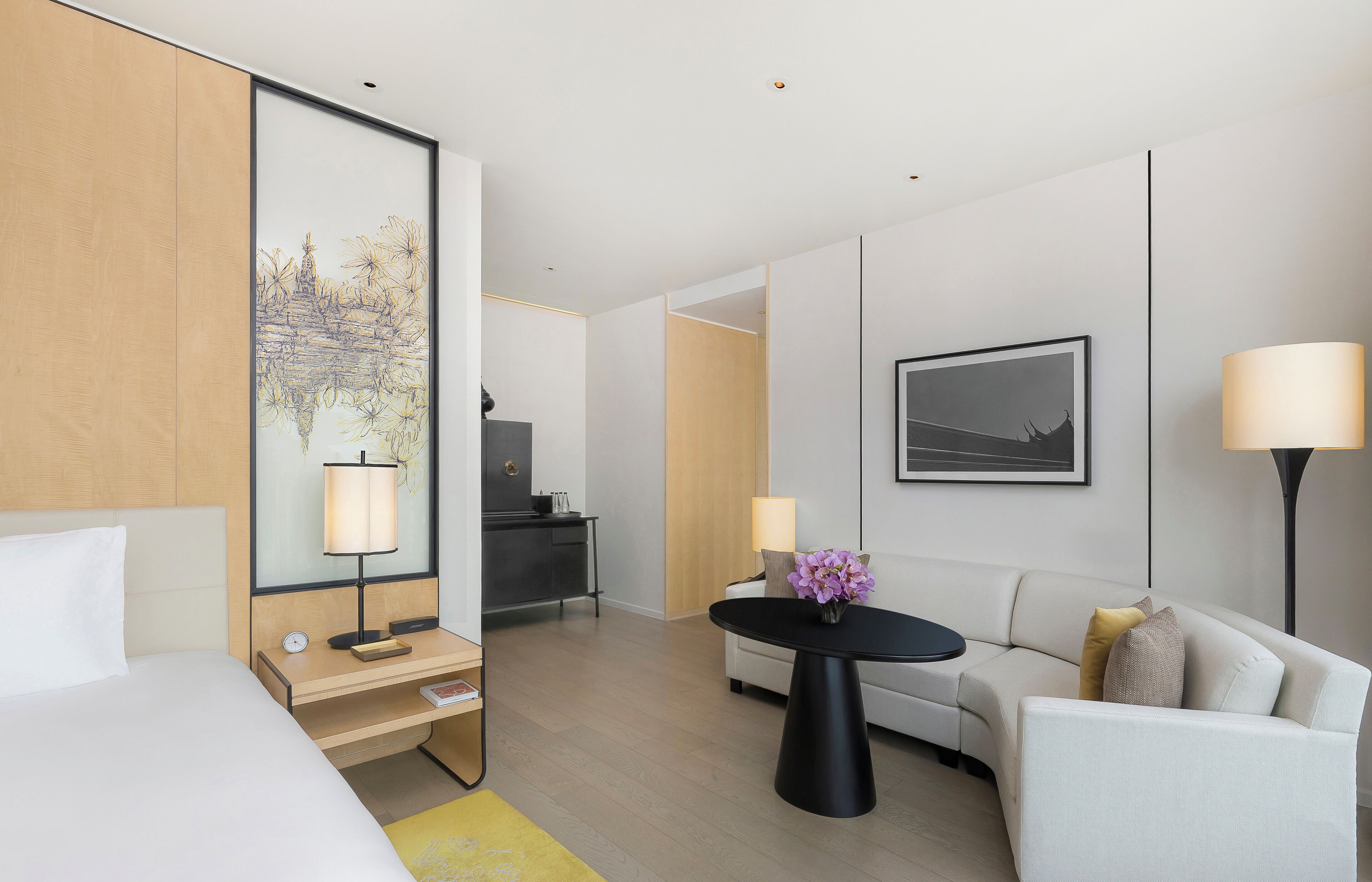 A modern hotel room with a bed, a desk with a lamp, a sofa, a coffee table with flowers, a desk lamp, and a framed picture on the wall.
