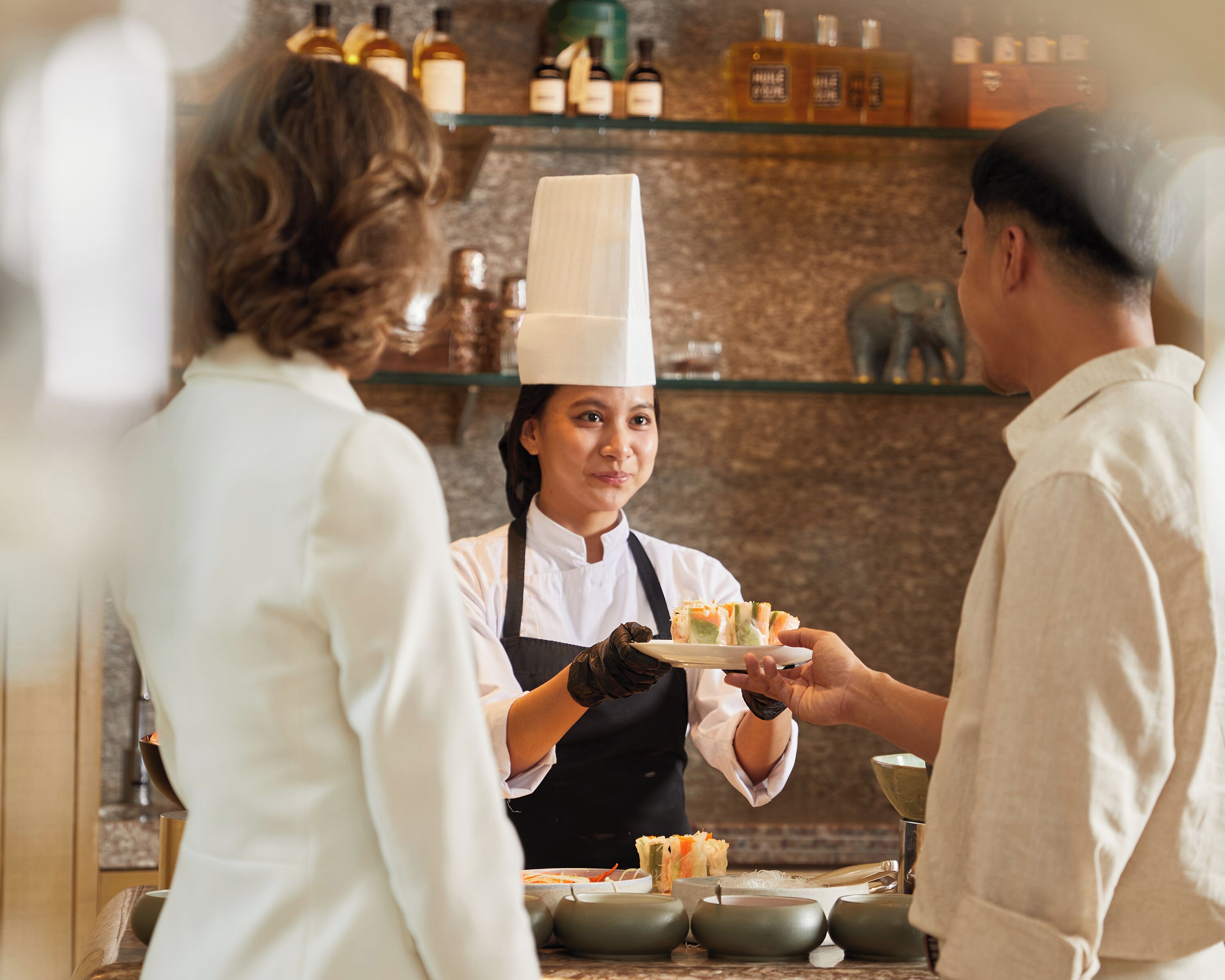 A chef serving sushi to a customer in a restaurant with a marble countertop and shelves of bottles in the background.