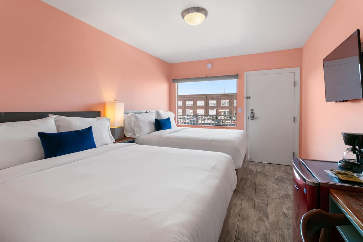 Quarto de hotel com duas camas, uma escrivaninha com cafeteira, televisão e janela com vista para a cidade.