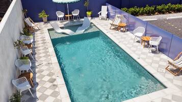 Piscina com piso quadriculado, cercada por cadeiras e mesas brancas, e uma parede azul.