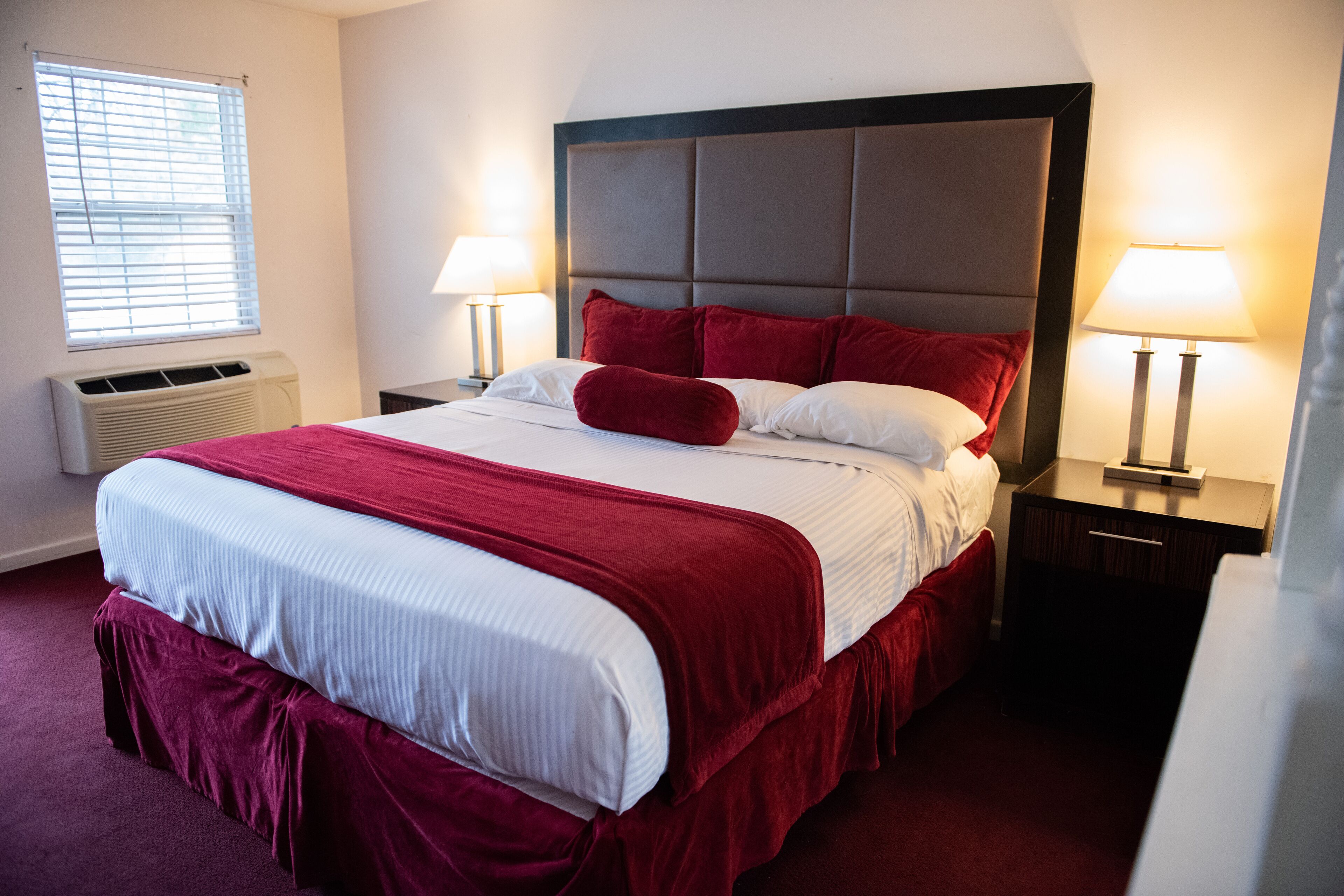 A neatly made bed with red and white bedding, two bedside lamps, and a window with blinds.
