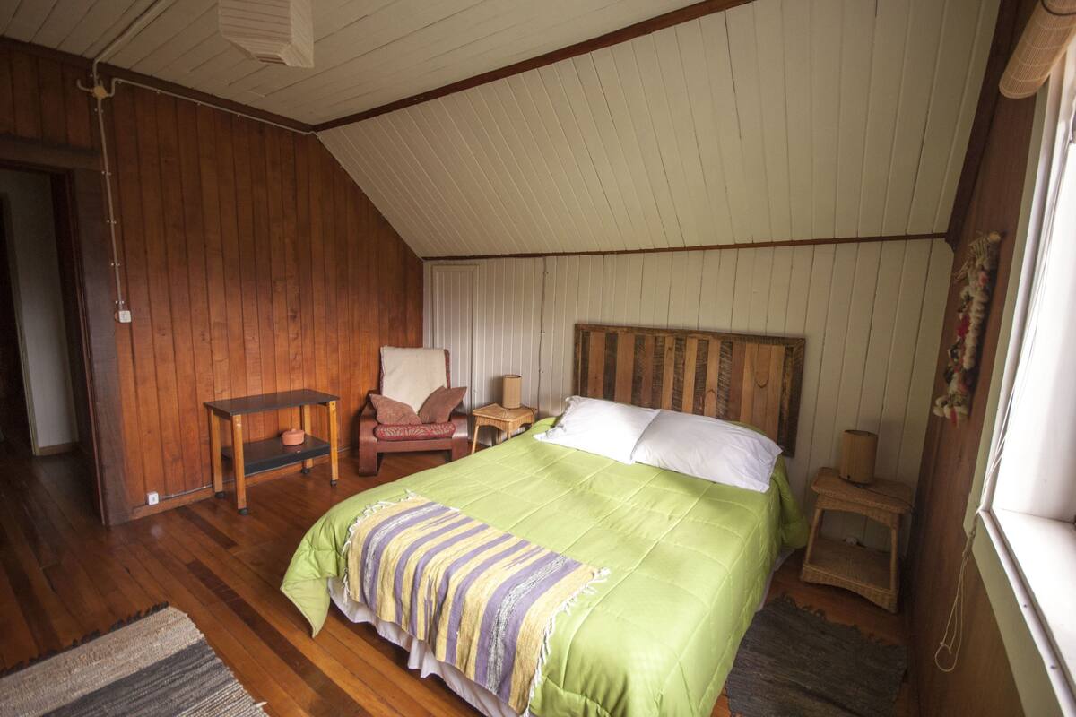 Ein Schlafzimmer mit Holzbalkendecke, einem Bett mit grüner Bettwäsche, einem Sessel, einem kleinen Tisch und einem Fenster.