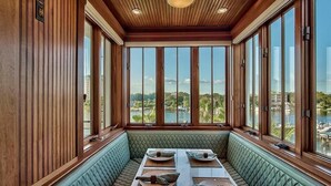 A dining area with a view of a lake and buildings, featuring wooden paneling, a green cushioned bench, and a table set with plates and napkins.