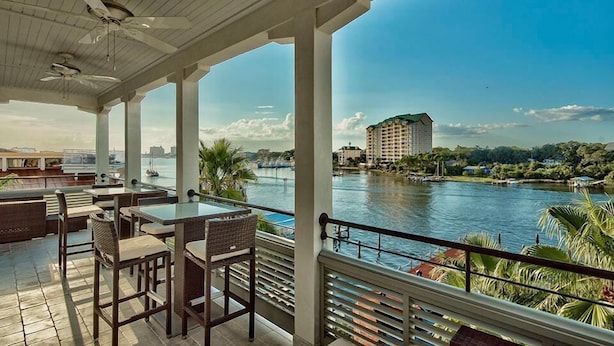 A covered outdoor seating area with tables and chairs, overlooking a river with buildings in the background.