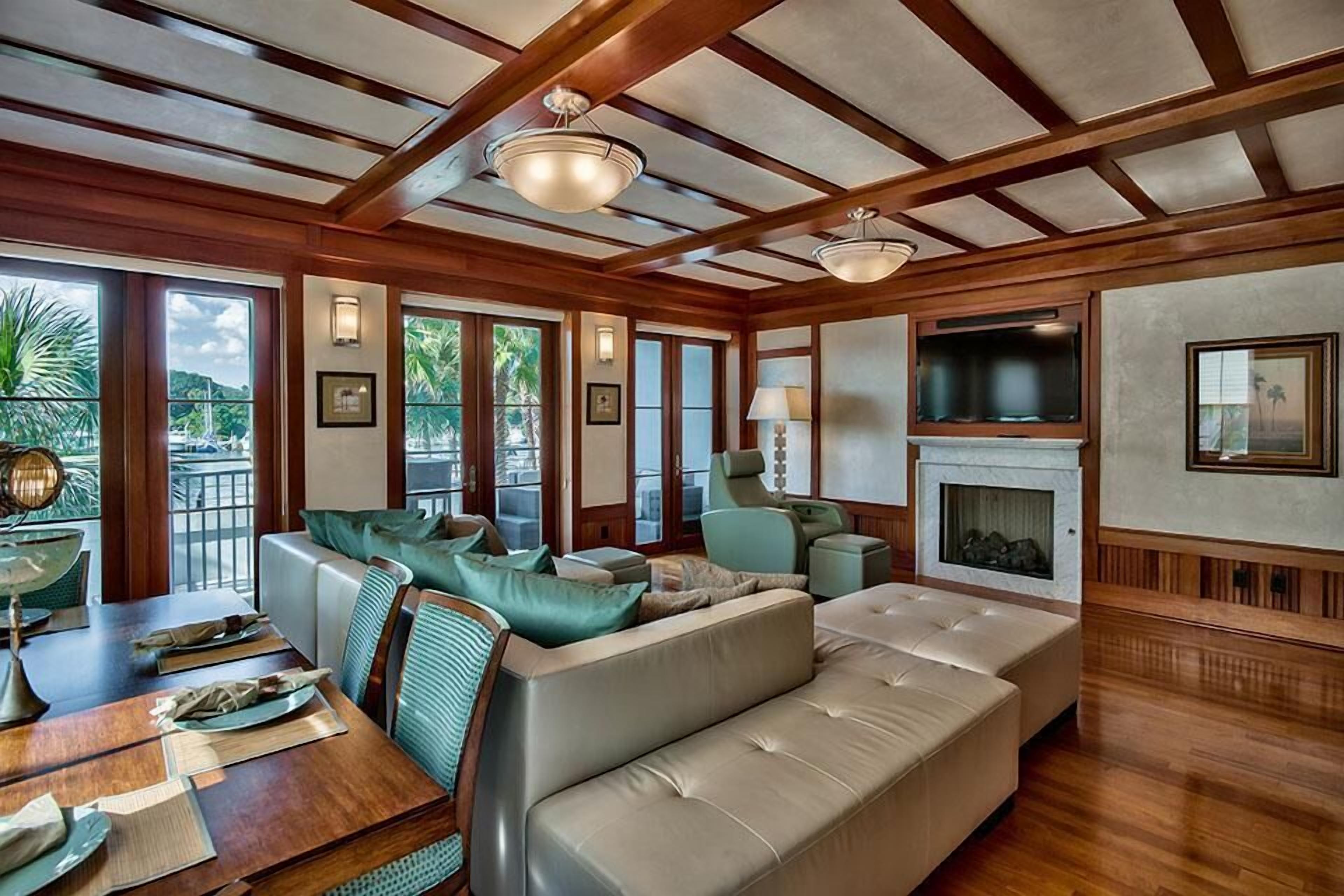 A spacious living room with a wooden ceiling, comfortable seating, a fireplace, and large windows offering a view of the outdoors.