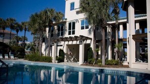 Un área junto a la piscina con una piscina azul cristalina, palmeras y un edificio de dos pisos con balcones.