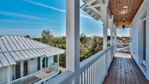 A covered porch with white railings and a wooden ceiling, overlooking a house with a metal roof and a view of the surrounding landscape.