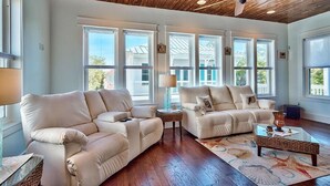A spacious living room with two beige sofas, a glass coffee table, and a wooden ceiling with a ceiling fan.