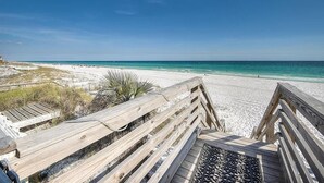 En träpromenad som leder till en sandstrand med klart, blått vatten.