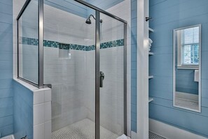 Een douche met glazen wanden, betegeld met witte tegels en een blauwe accentrand, een witte wastafel en een spiegel met een witte omlijsting.