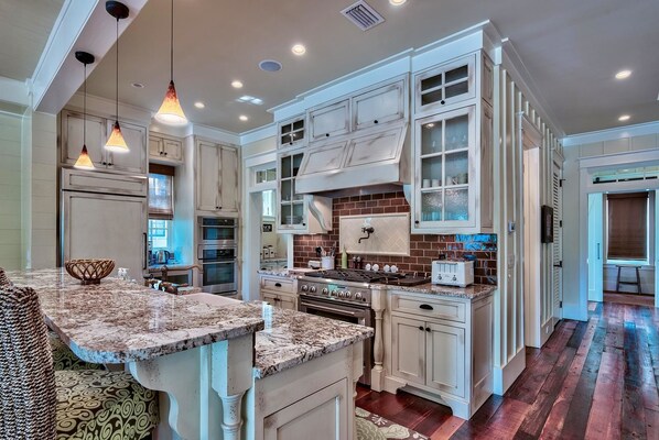 A spacious kitchen with granite countertops, white cabinetry, and a large island. There are built-in ovens and a range with a backsplash.
