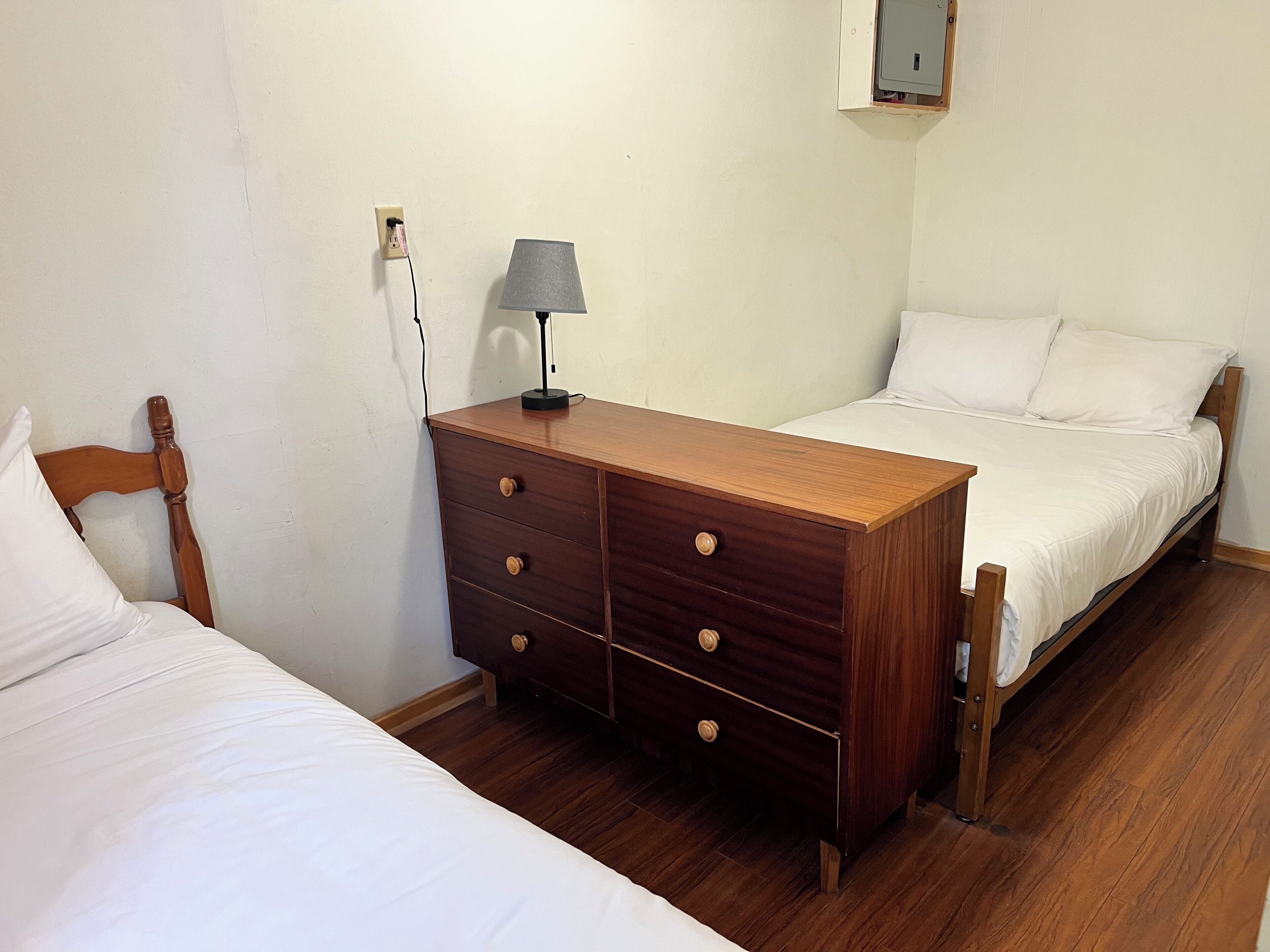 A bedroom with two beds, a wooden dresser, a lamp, and a wall socket.