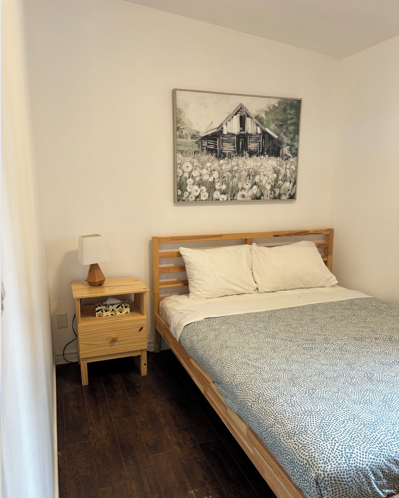 A bedroom with a wooden bed, a nightstand with a lamp, a small drawer, and a painting of a barn on the wall.