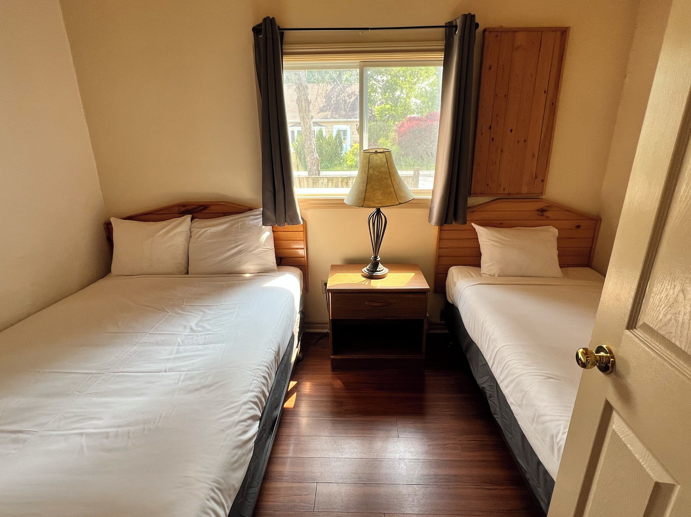 A room with two single beds, a wooden headboard, a nightstand with a lamp, and a window with curtains.